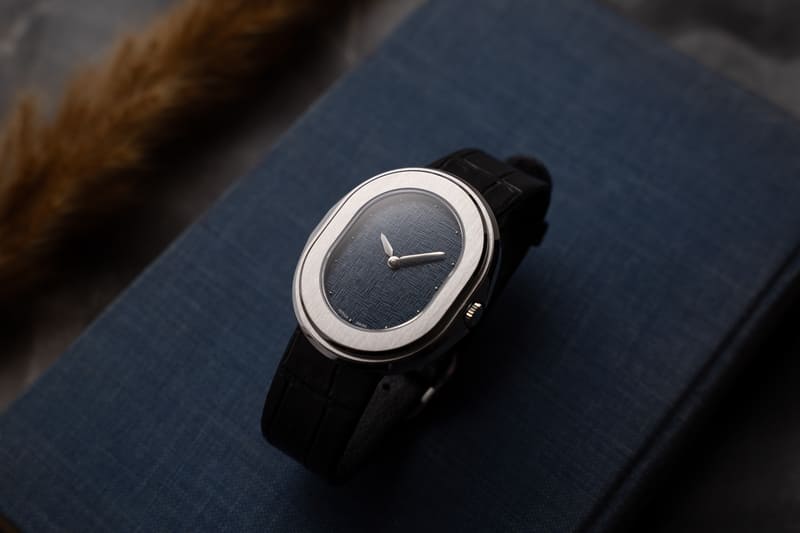 Luxury stainless steel oval Serica Parade 1174 dress watch with blue linen textured dial and black leather strap, minimalist Swiss wristwatch close-up on elegant fabric background